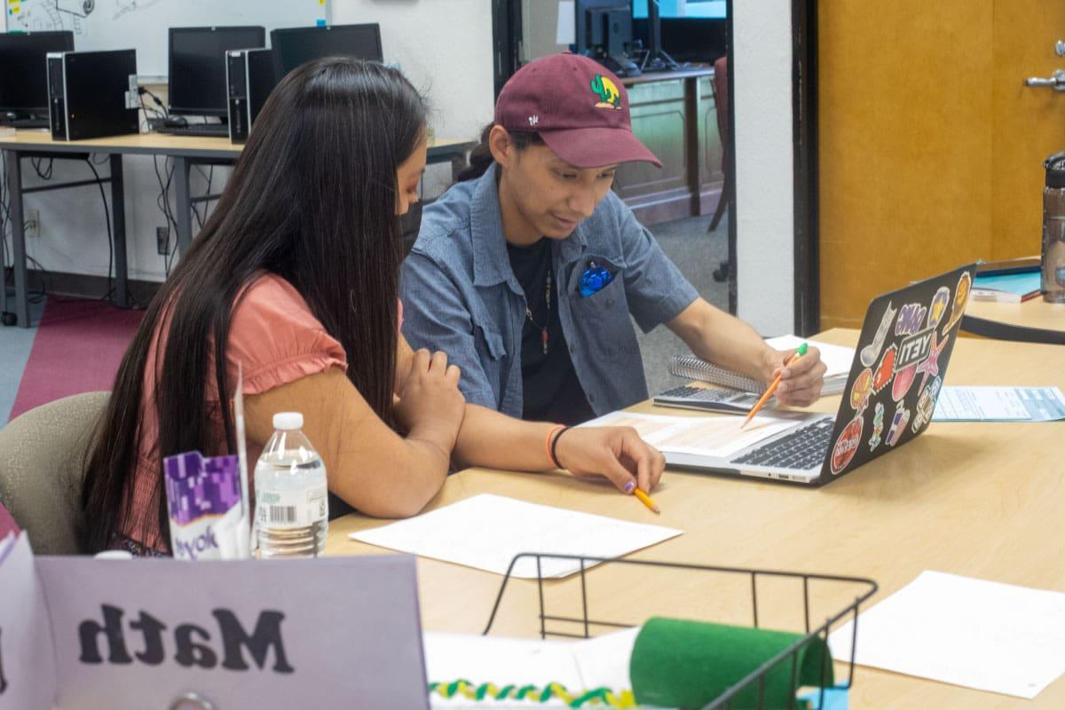 Two individuals sit at a table with an open laptop in front of them, reviewing text on a piece of paper